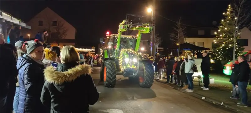 Освещенный зеленый трактор проезжает через деревню, а люди стоят на обочине дороги