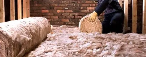 Рабочий разматывает изоляцию из стекловаты на чердаке.